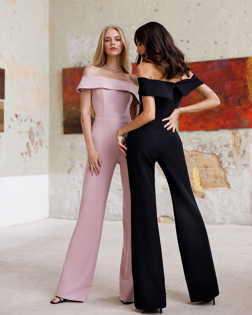 Two women wearing off-shoulder jumpsuits standing in a room with a textured wall.
