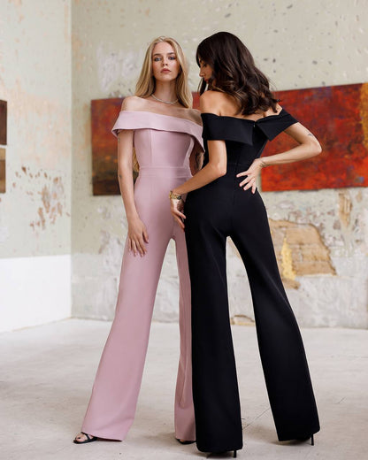 Two women wearing off-shoulder jumpsuits standing in a room with a textured wall.