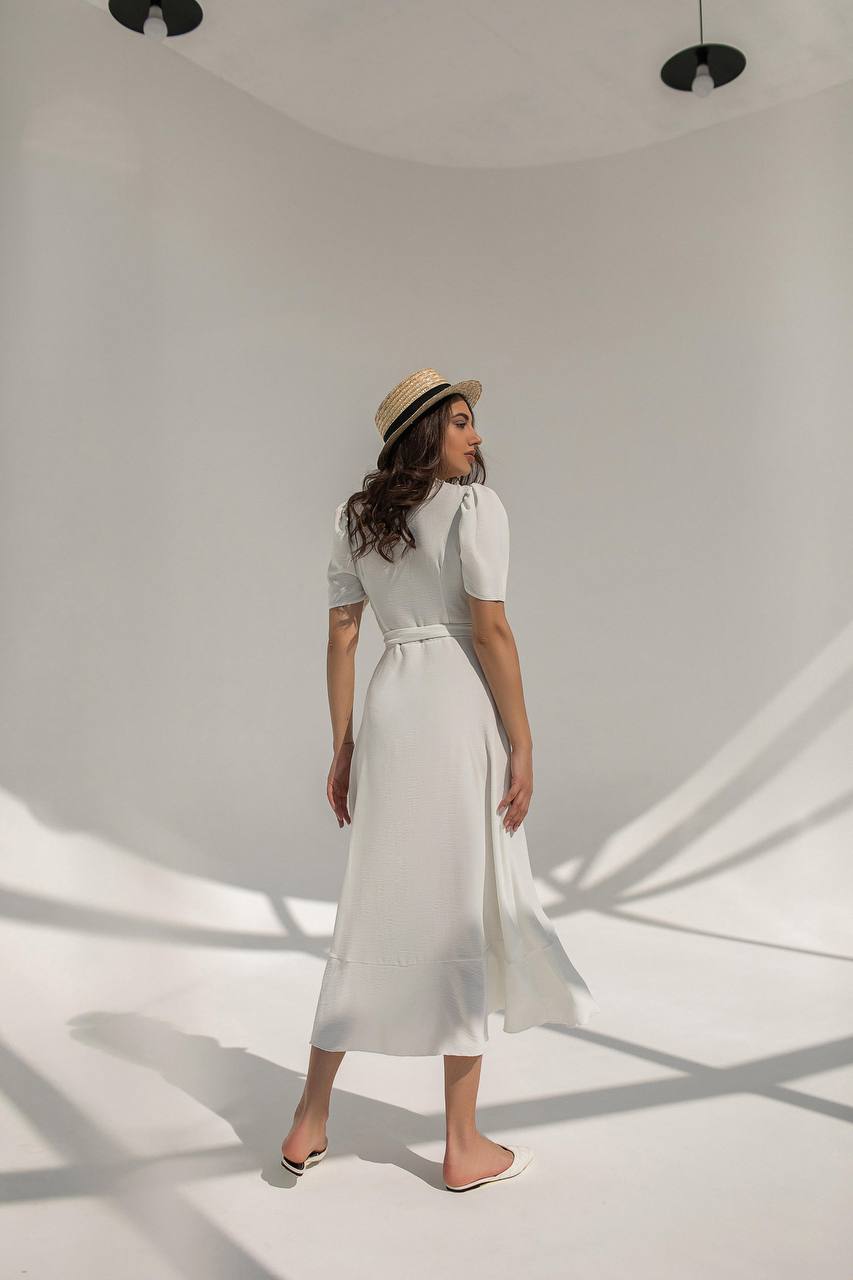 Woman in a white dress and straw hat standing in a softly lit room with shadows on the wall.