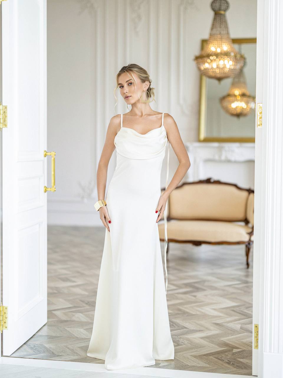 A woman modeling a floor-length white satin gown with thin spaghetti straps and a draped neckline, standing in an elegant room with a herringbone wood floor, a beige sofa, and a chandelier.