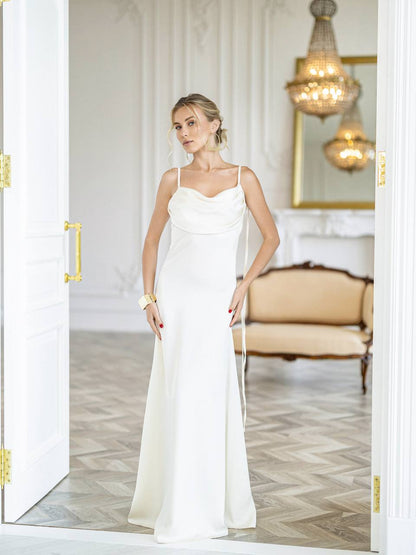 A woman modeling a floor-length white satin gown with thin spaghetti straps and a draped neckline, standing in an elegant room with a herringbone wood floor, a beige sofa, and a chandelier.