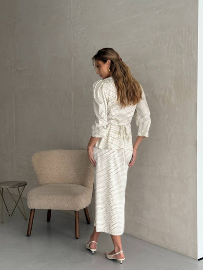 Woman in a white textured outfit standing in a minimalistic room with a chair and small table.
