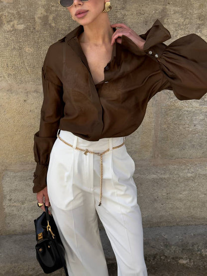 Person wearing a brown blouse and white pants against a stone wall.