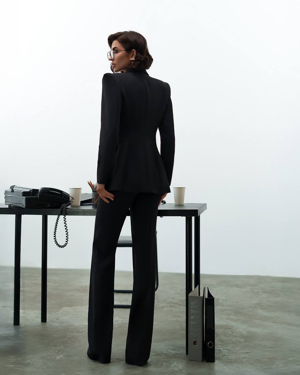 Person in a black suit standing in an office setting with a desk and phone.