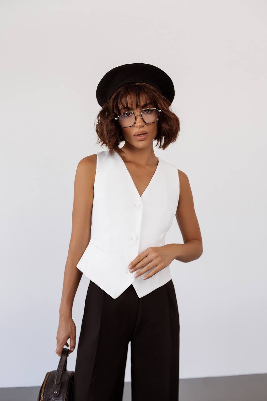 Person wearing a white sleeveless top and black pants with a black cap and glasses, holding a brown bag against a light gray background.