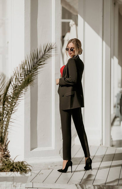 Woman in a black suit standing in front of a white building with a palm leaf in the foreground.