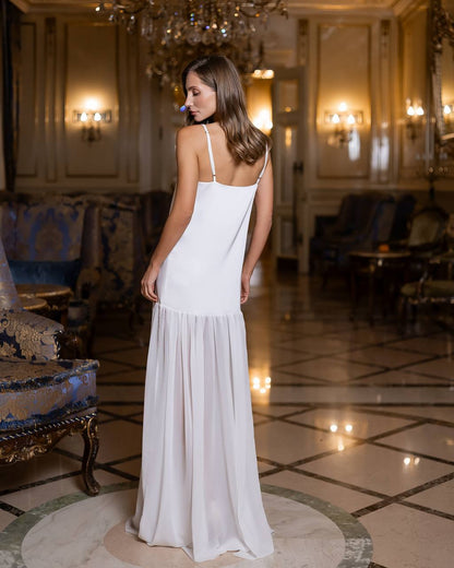 Woman in a white dress standing in an elegant room with chandeliers and decor.