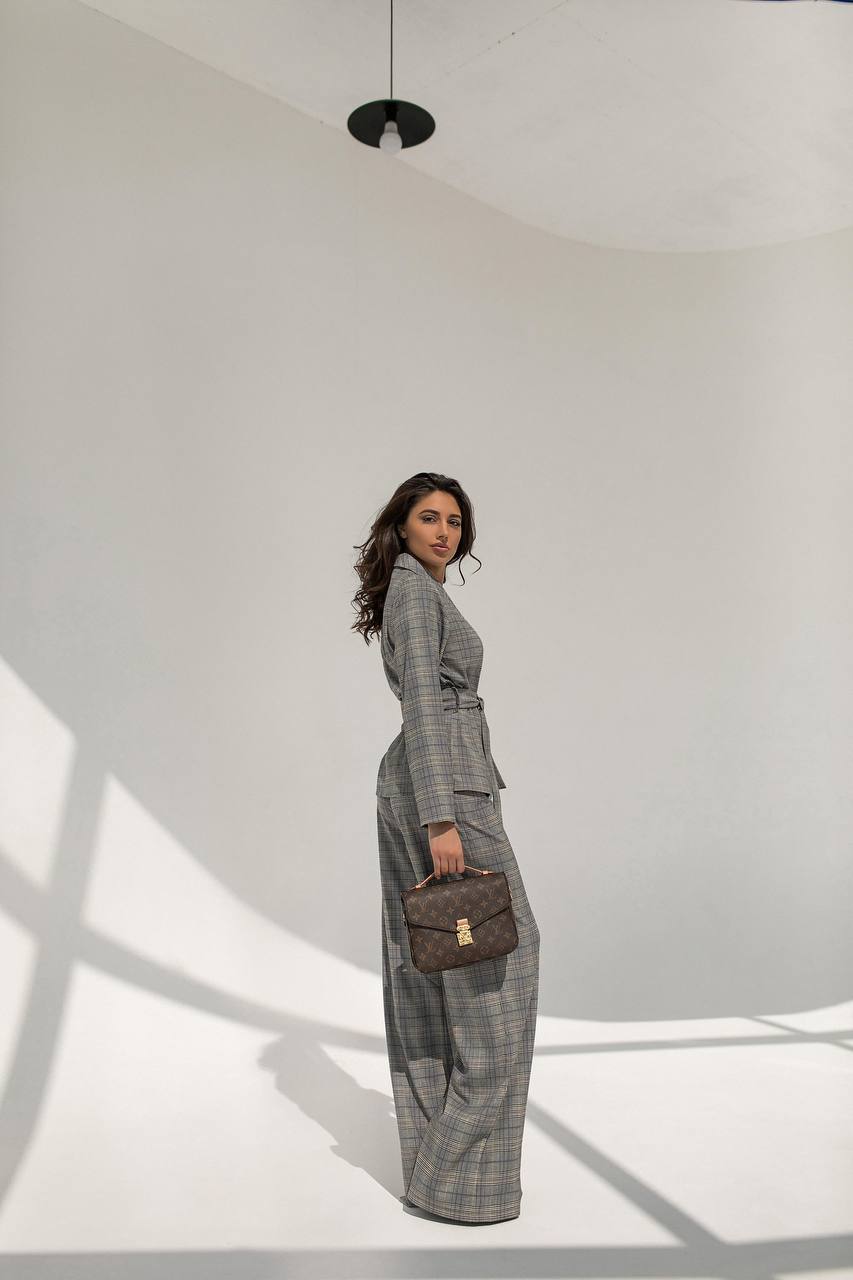 Woman in a gray checkered suit holding a brown handbag in a minimalistic setting