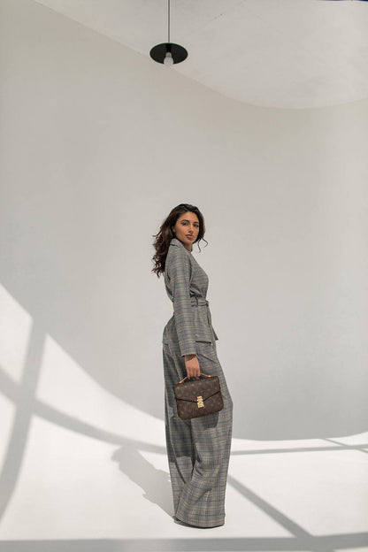 Woman in a gray checkered suit holding a brown handbag in a minimalistic setting