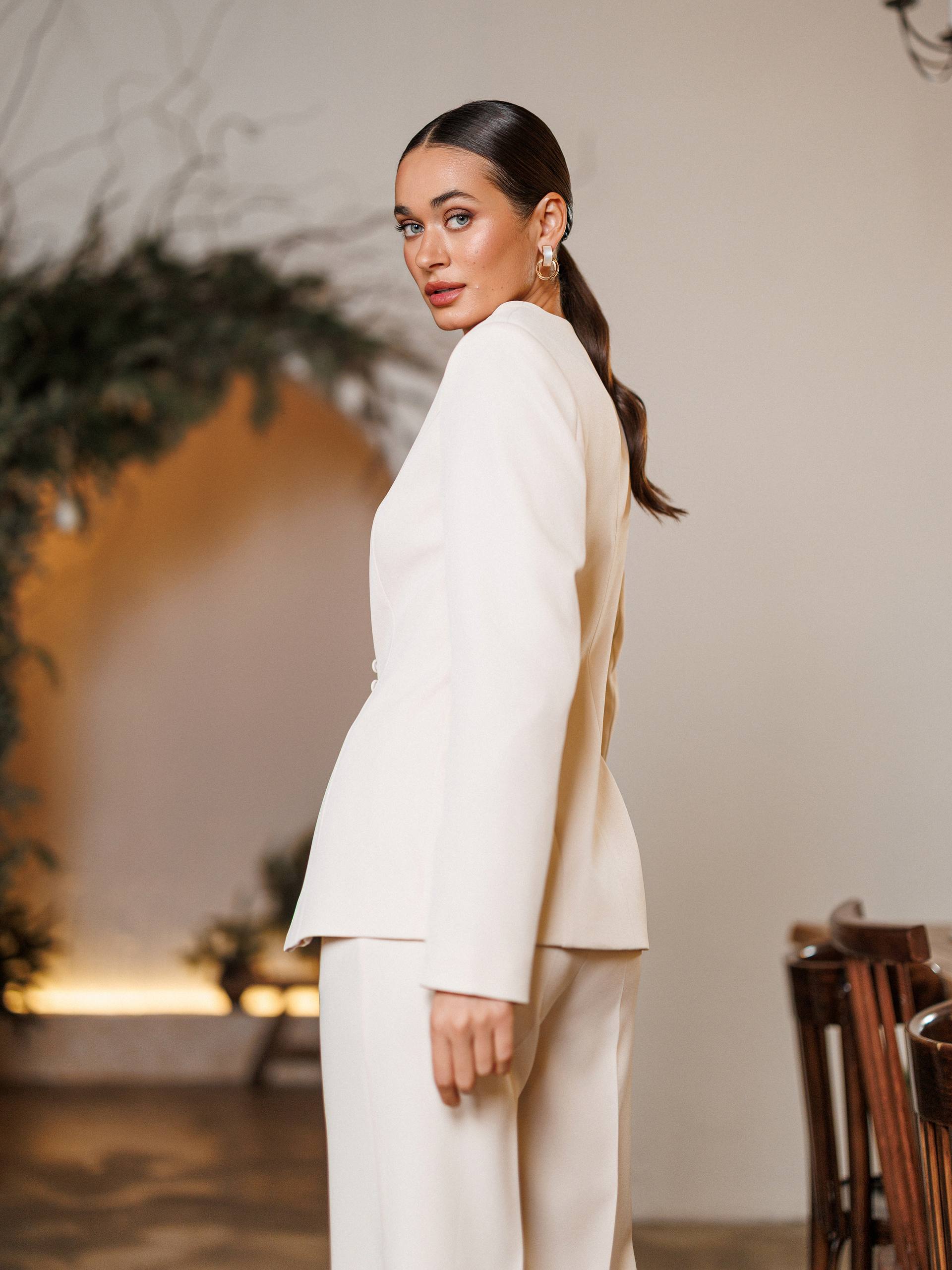Woman in a white suit standing in an indoor setting with decorative elements.
