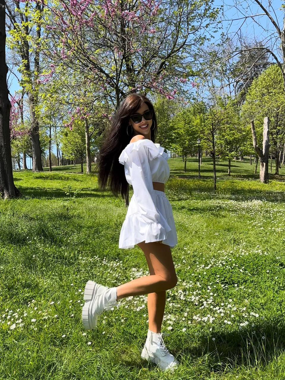 Woman in a white outfit standing in a park with green grass and trees.