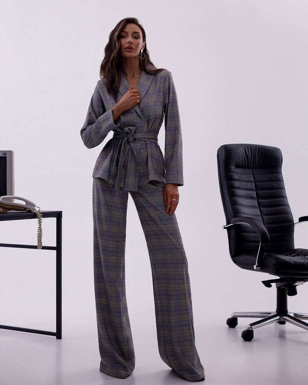Woman in a plaid suit standing in an office setting with a chair and desk.
