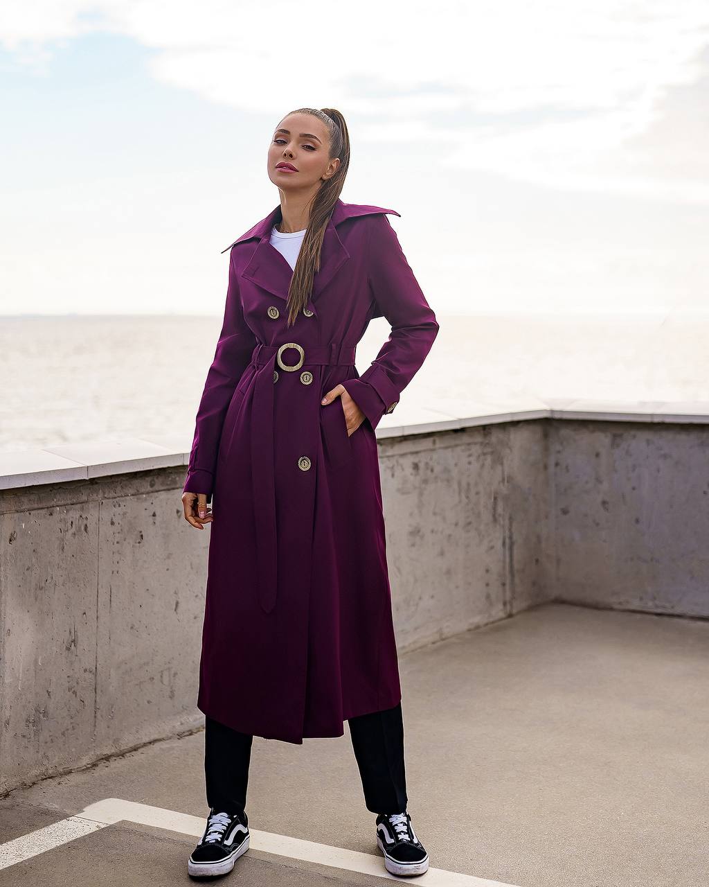 Woman wearing a purple trench coat on a rooftop with a concrete wall and sky background