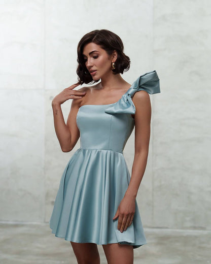 Woman wearing a light blue one-shoulder dress with a bow detail against a plain background