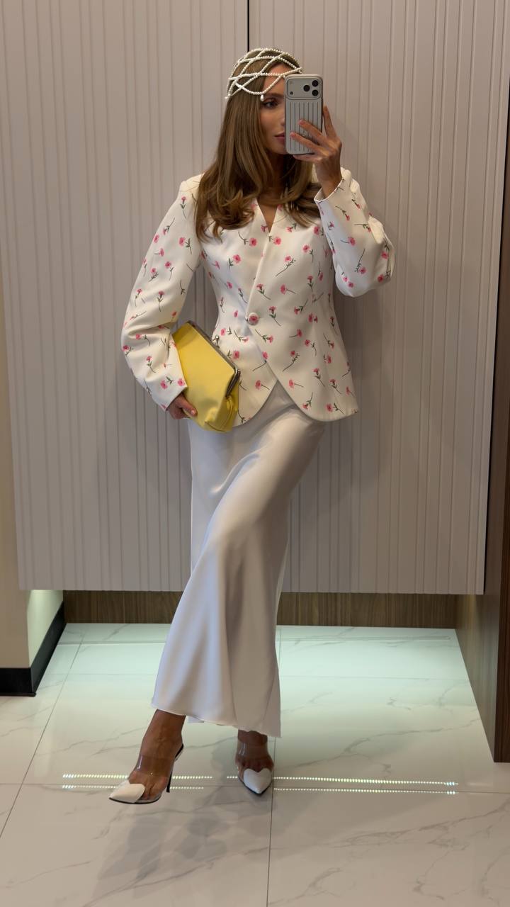 Person wearing a floral blouse and white pants, standing in a room with a neutral background.