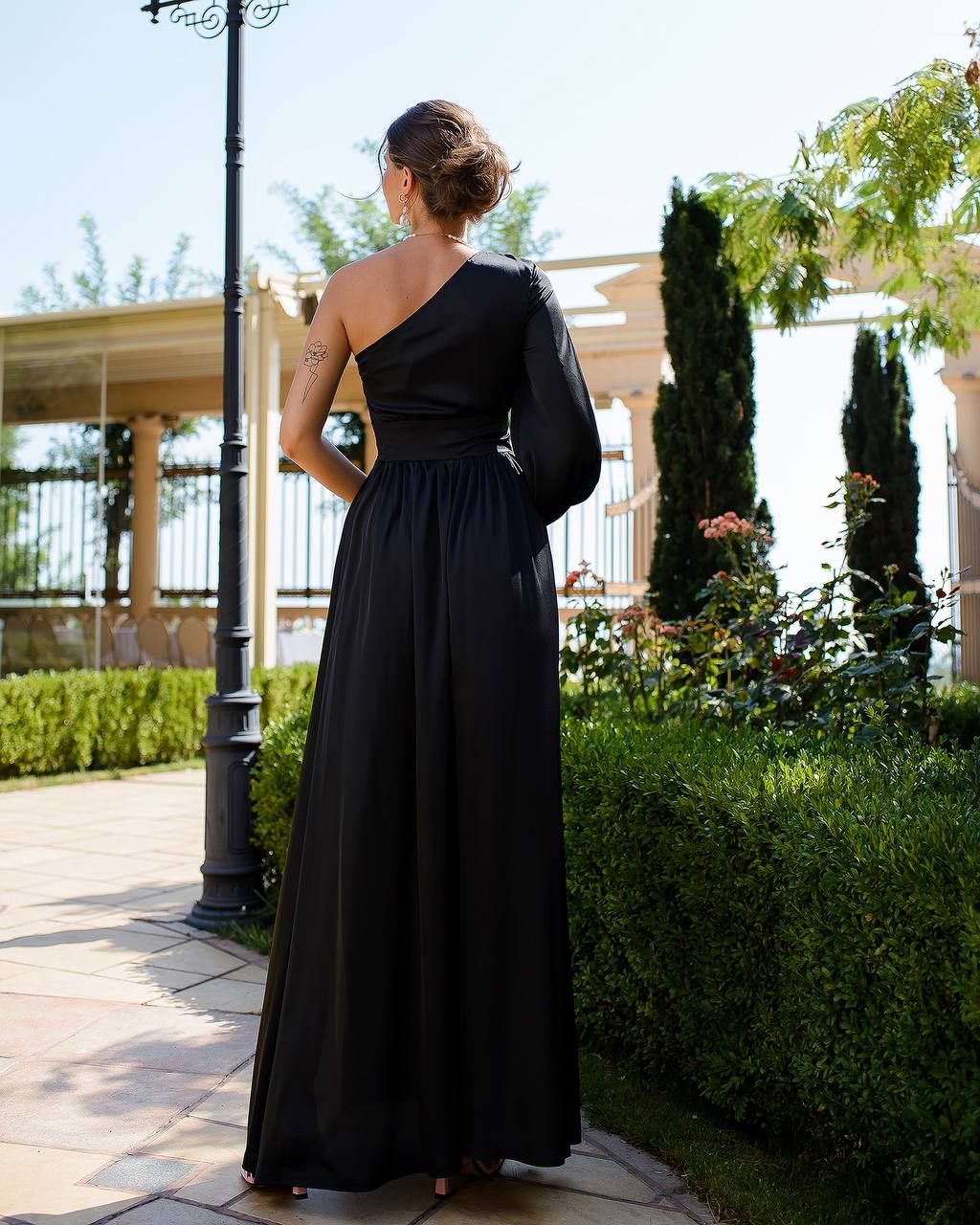Woman in a black one-shoulder dress standing outdoors with greenery and a building in the background.
