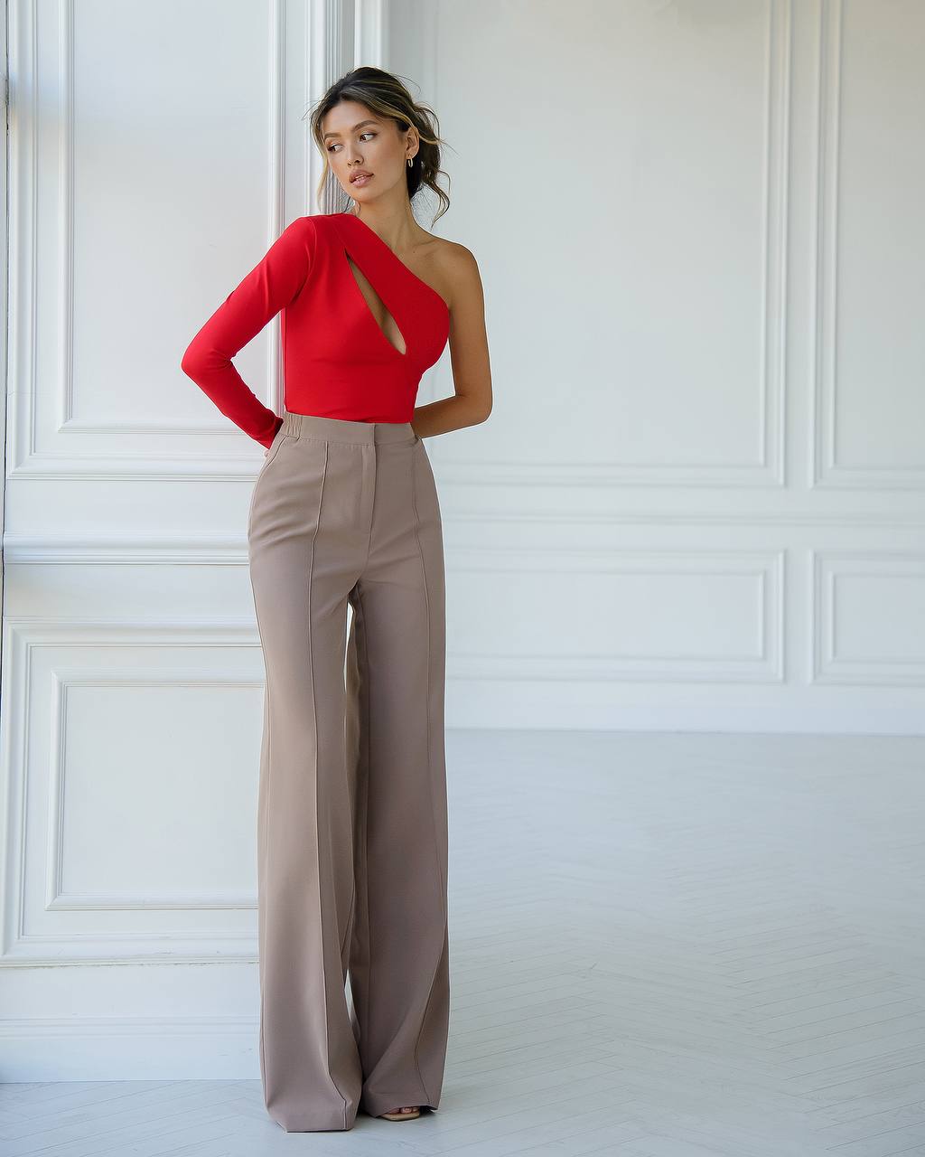 Woman wearing a red one-shoulder top and beige pants against a white paneled wall.