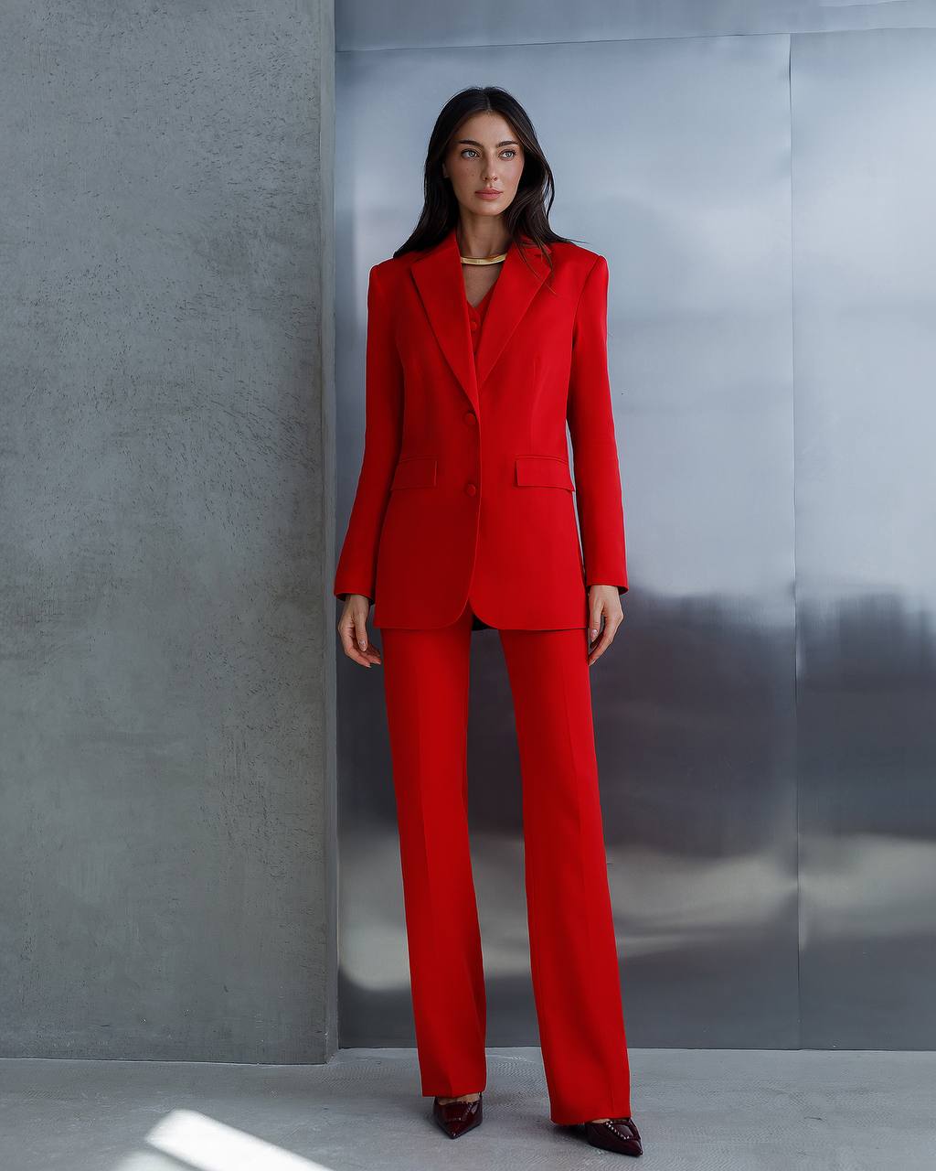 Woman wearing a red suit standing against a gray wall.