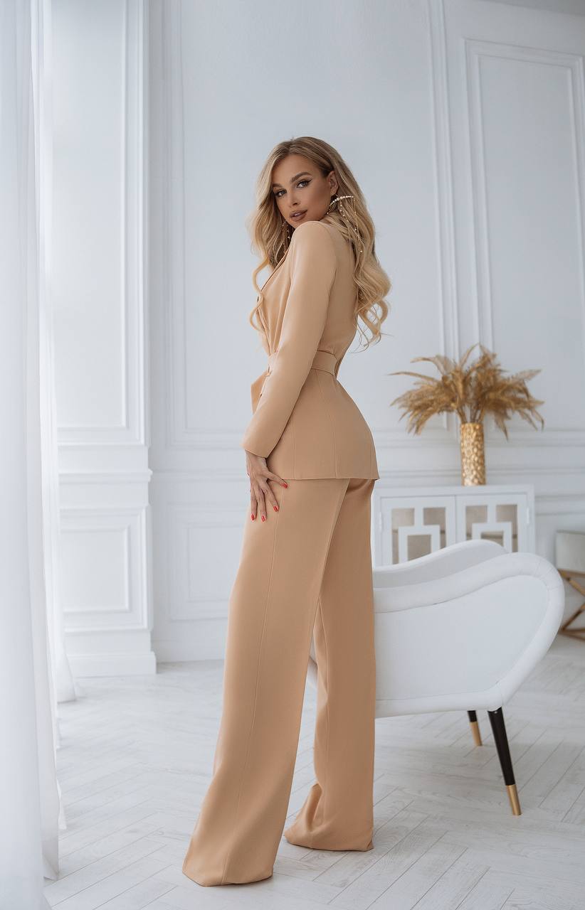 Woman in a beige outfit standing in a stylish room with white walls and decor.