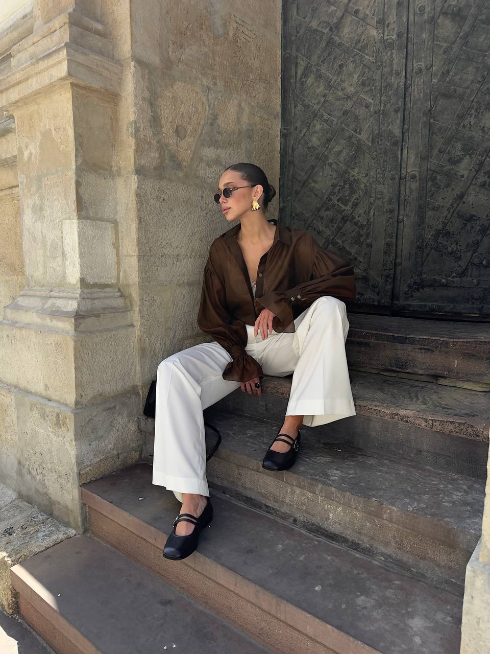 Woman sitting on stone steps wearing a brown blouse and white pants.