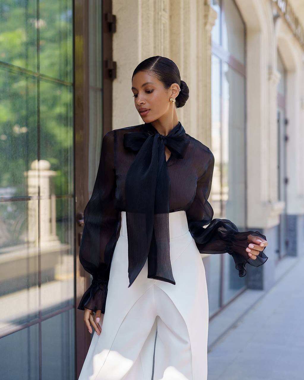 Woman wearing a black blouse with a large bow and white pants standing outdoors.