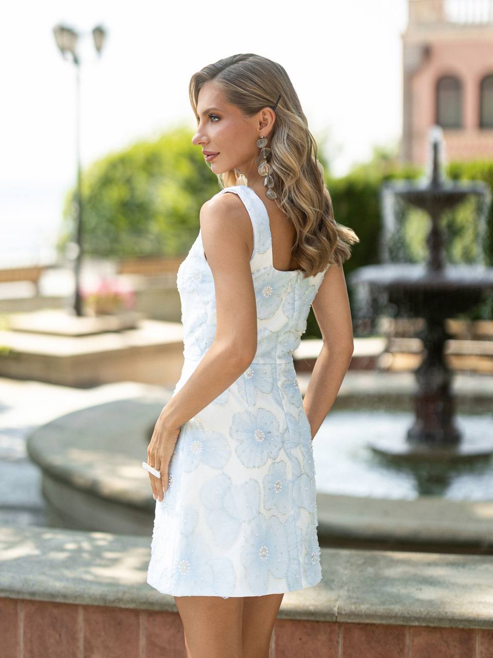 A woman wearing a sleeveless white dress with light blue floral applique, standing outdoors in front of a fountain and greenery.