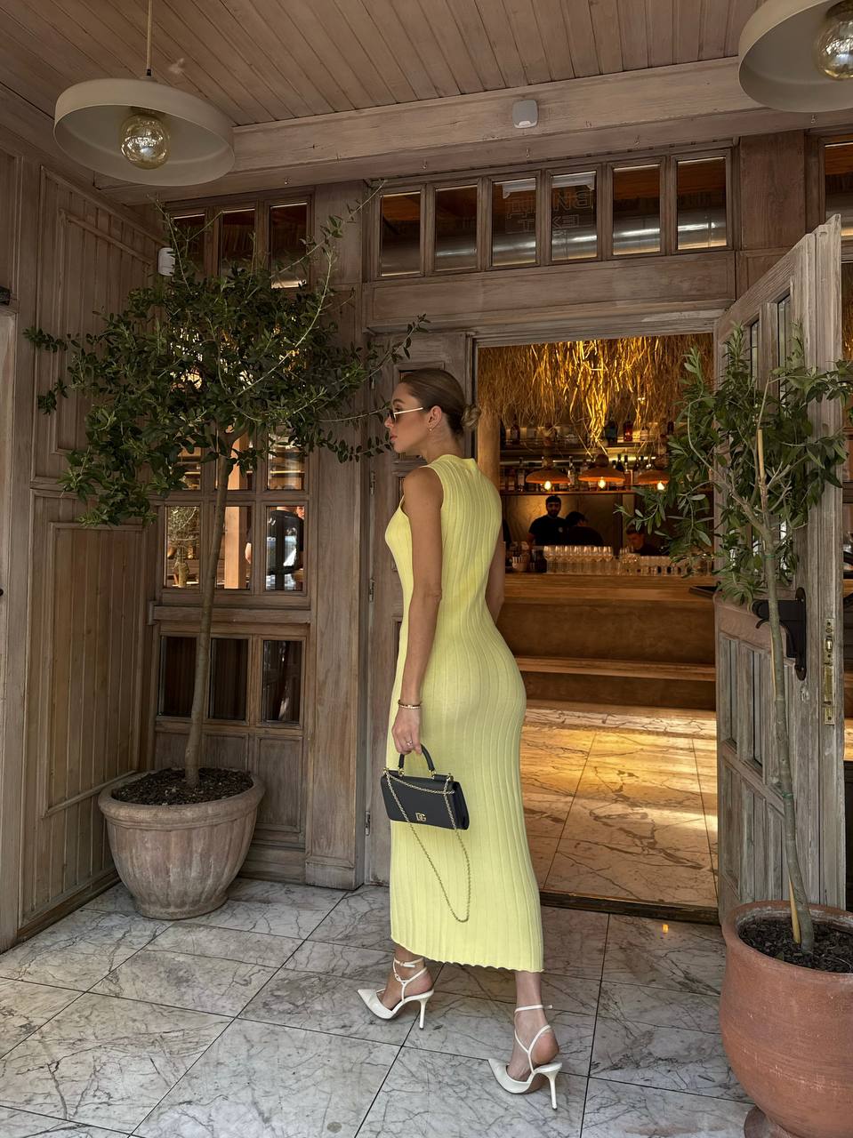 Woman in a yellow dress standing in a stylish interior setting with plants and wooden decor.