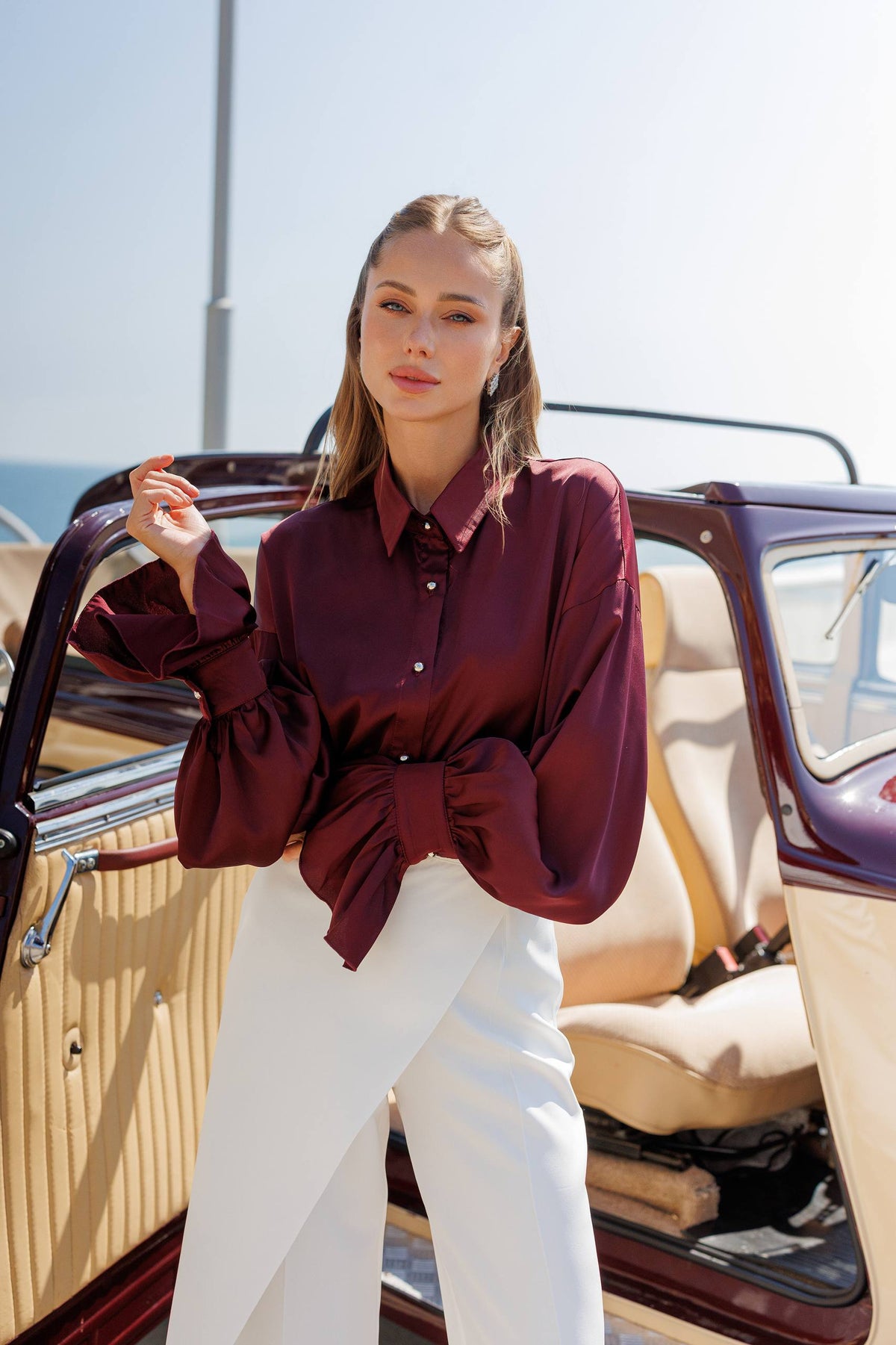 Woman in a burgundy shirt and white pants standing next to a vintage car.