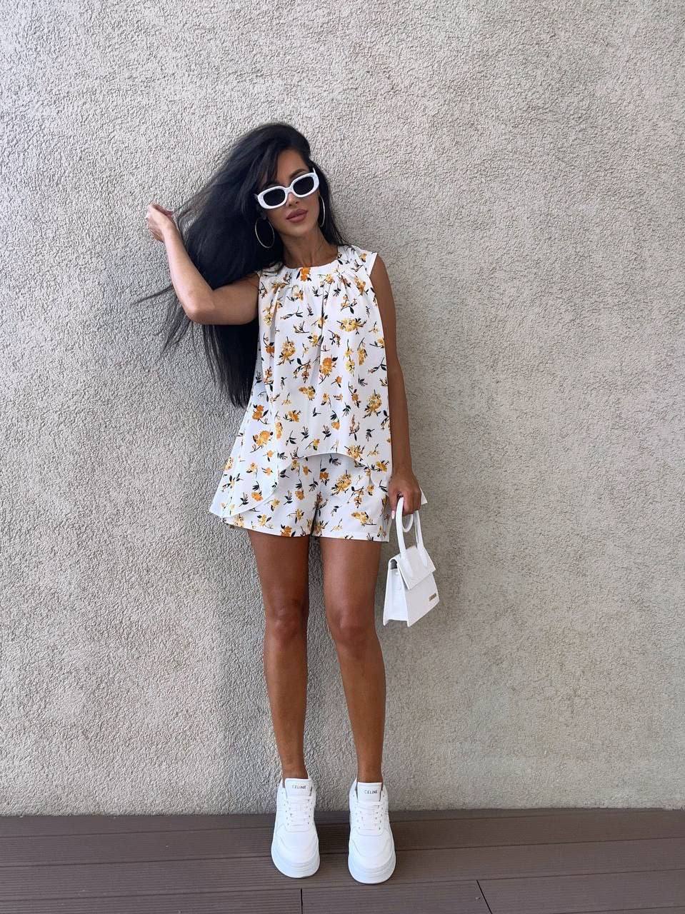 Woman wearing a floral dress and white sneakers against a plain wall.