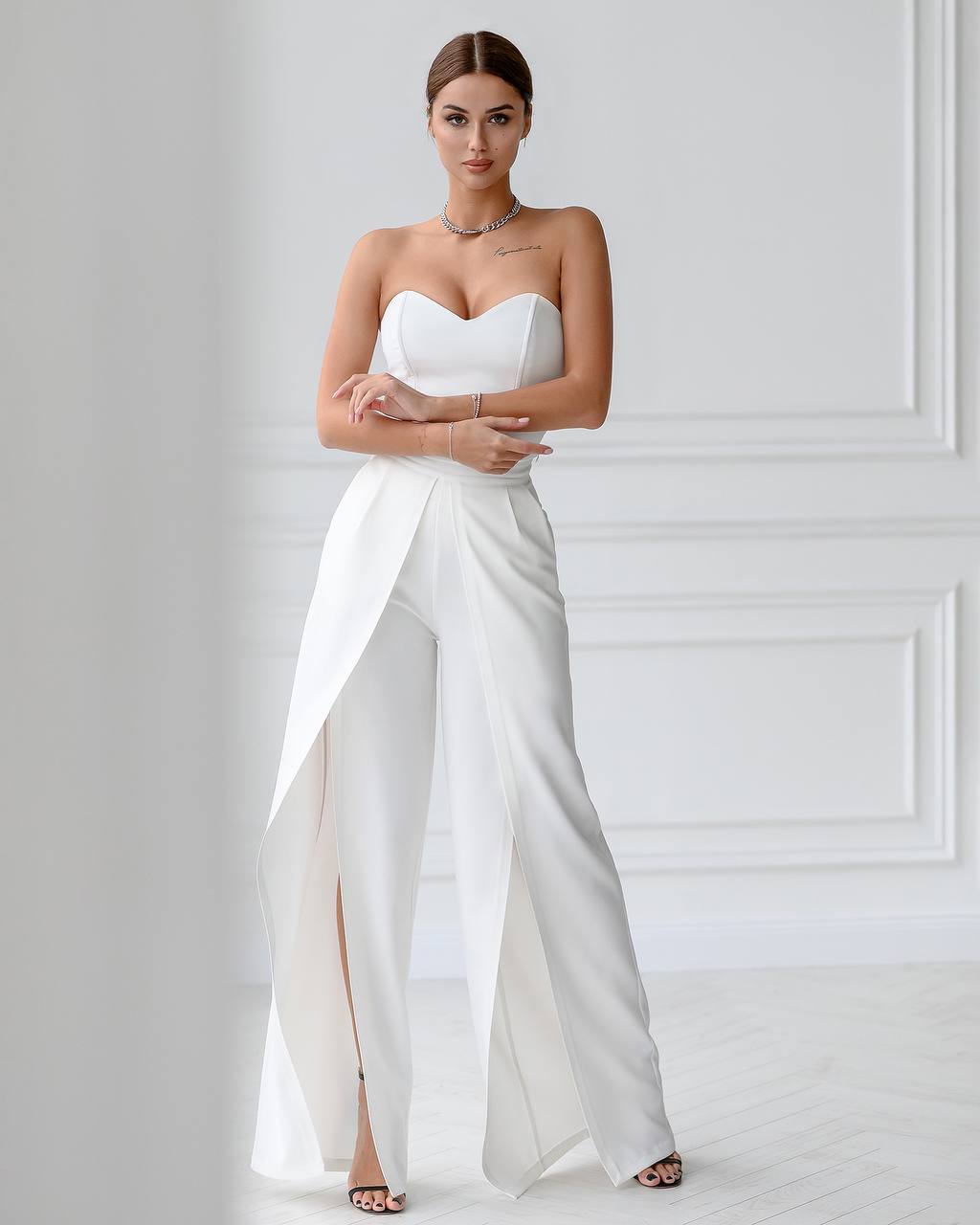 Woman wearing a white strapless jumpsuit with a high slit on a plain background