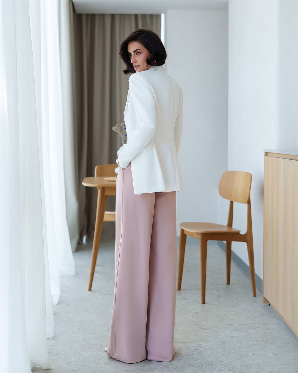 Woman in a white blazer and pink pants standing in a modern room with wooden furniture.