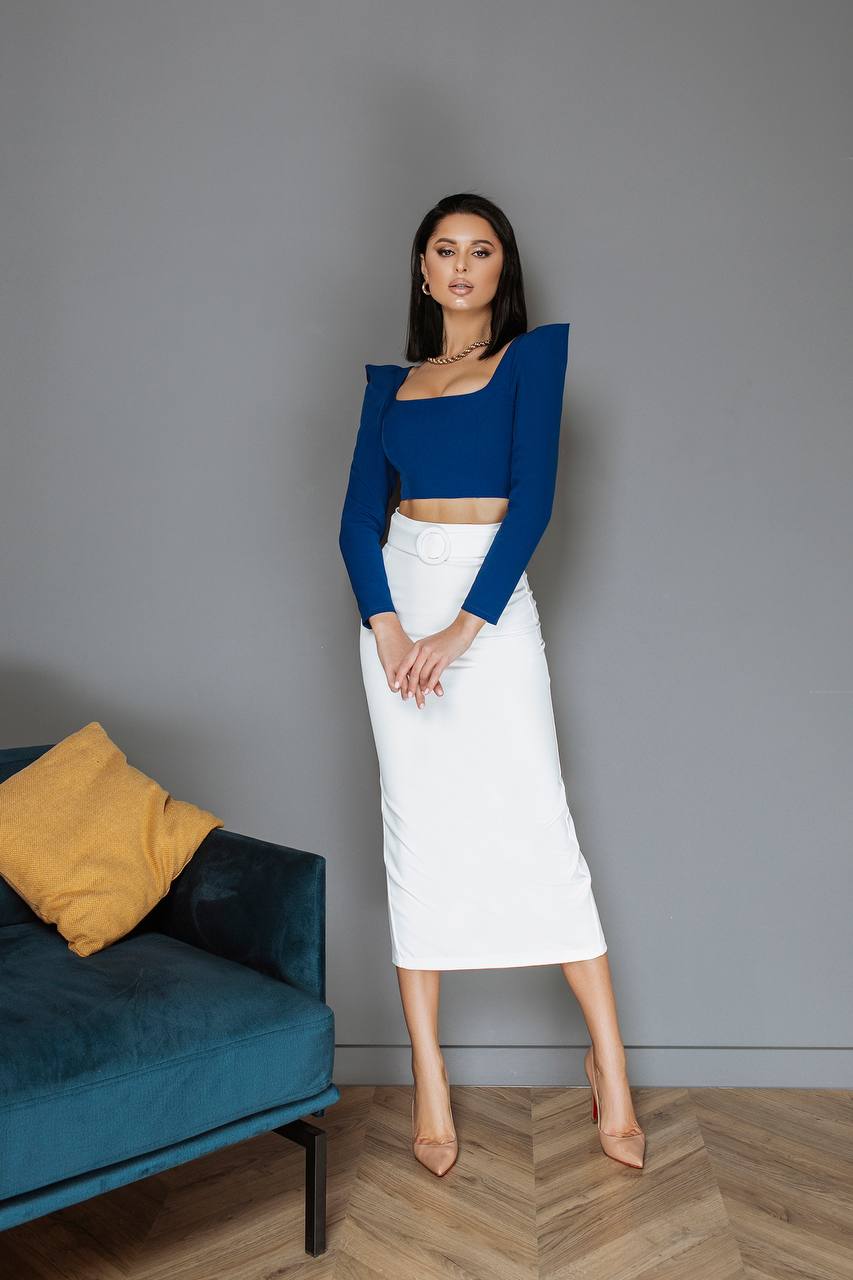 Woman in a blue top and white skirt standing in a room with a gray wall and blue couch.