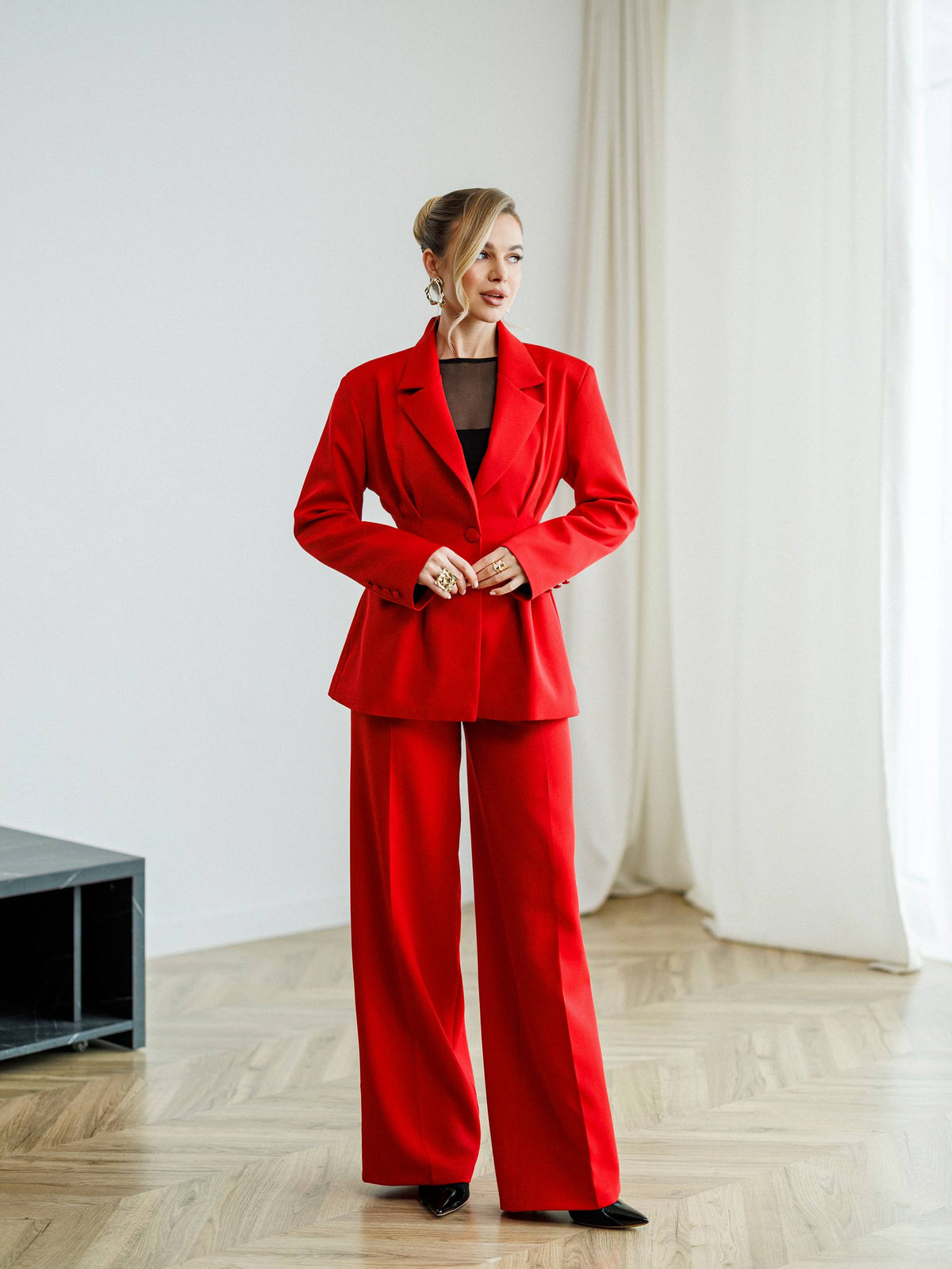 A woman models a bright red two-piece suit consisting of a double-breasted blazer and matching wide-leg trousers, styled over a black top in a minimalist room with light wood floors and white curtains.