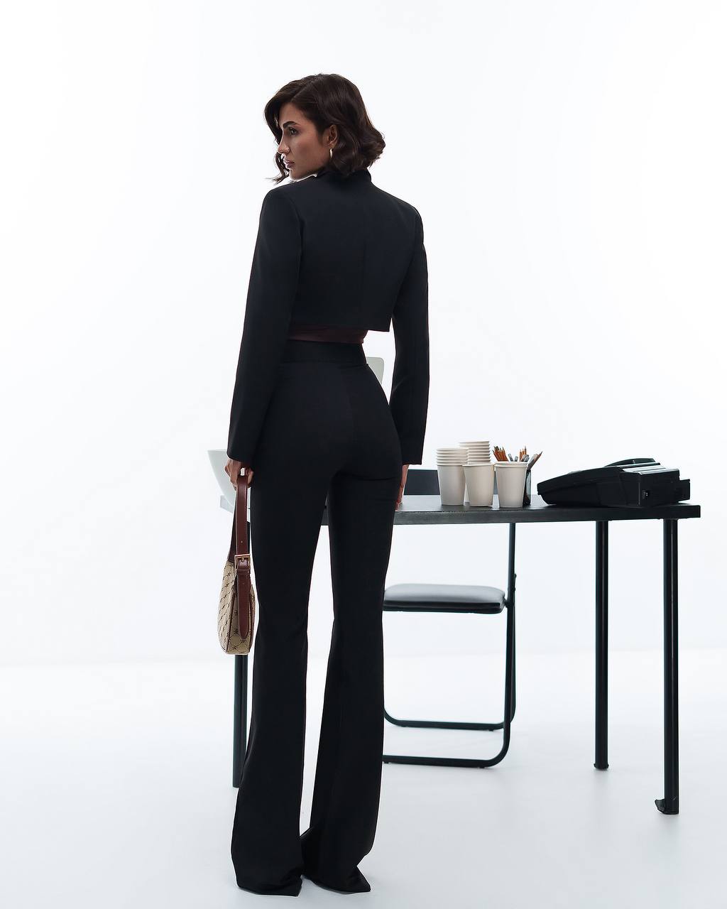 Woman in a black suit standing in an office setting with a desk and chair.