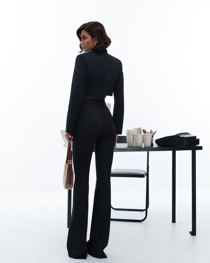 Woman in a black suit standing in an office setting with a desk and chair.