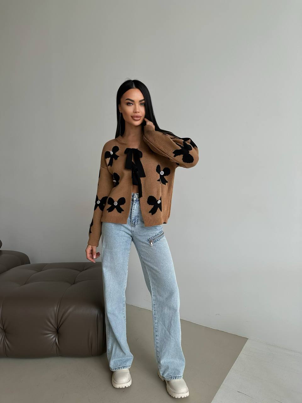 Person wearing a brown sweater with black floral patterns and light blue jeans indoors.