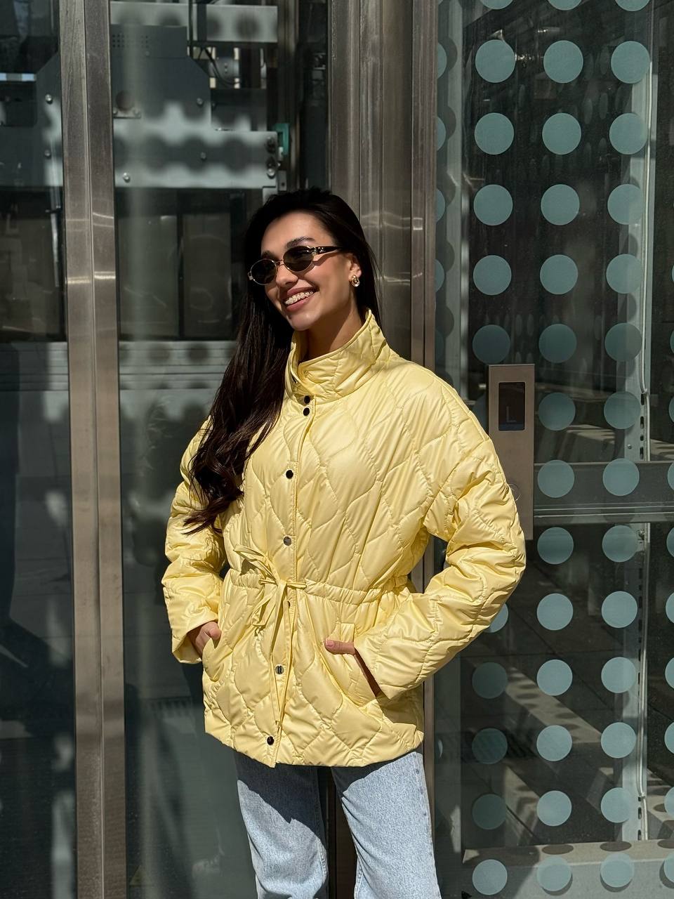 Woman wearing a yellow quilted jacket standing in front of a glass wall with circular patterns.