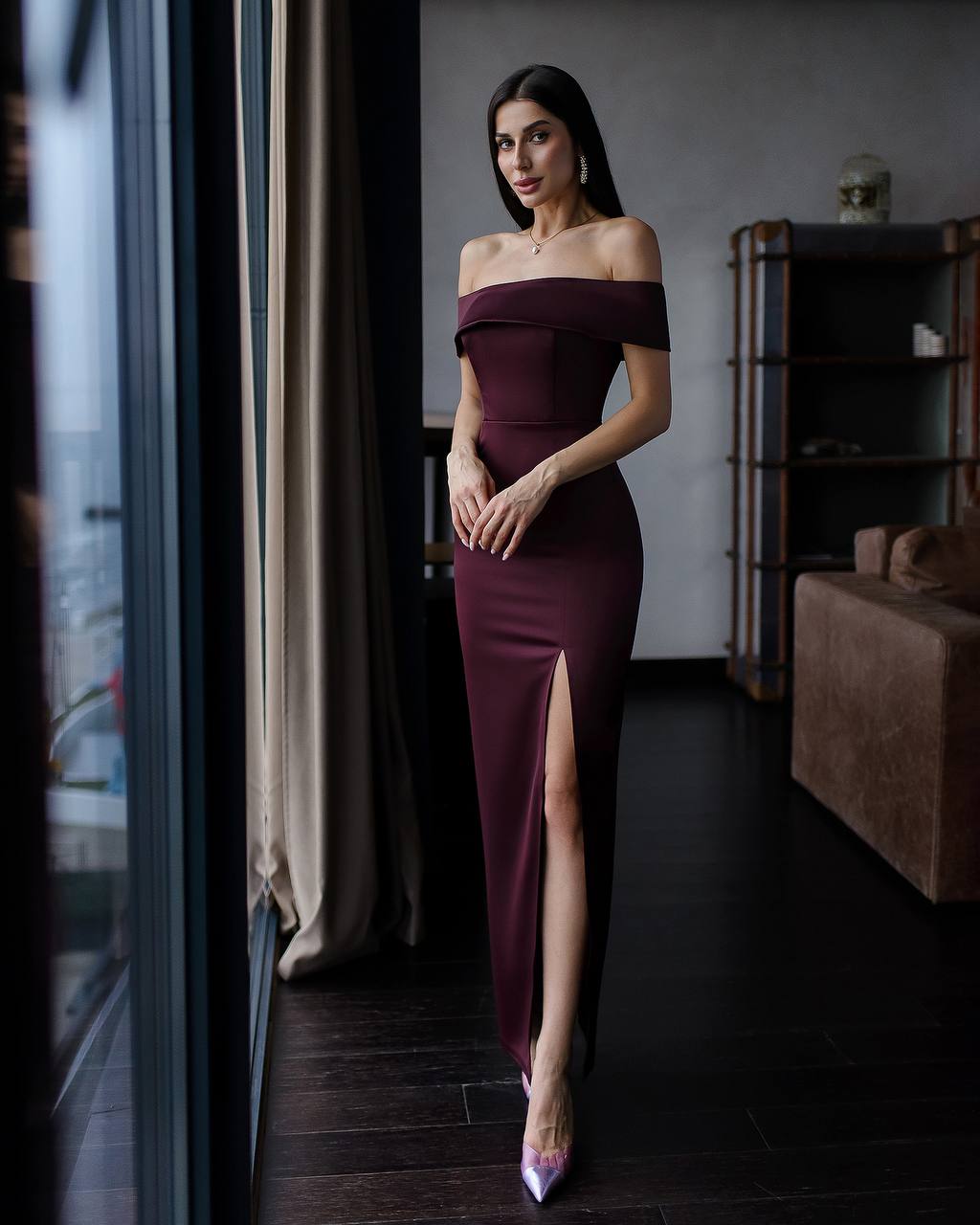 Woman in a burgundy off-shoulder dress with a slit standing in a room.