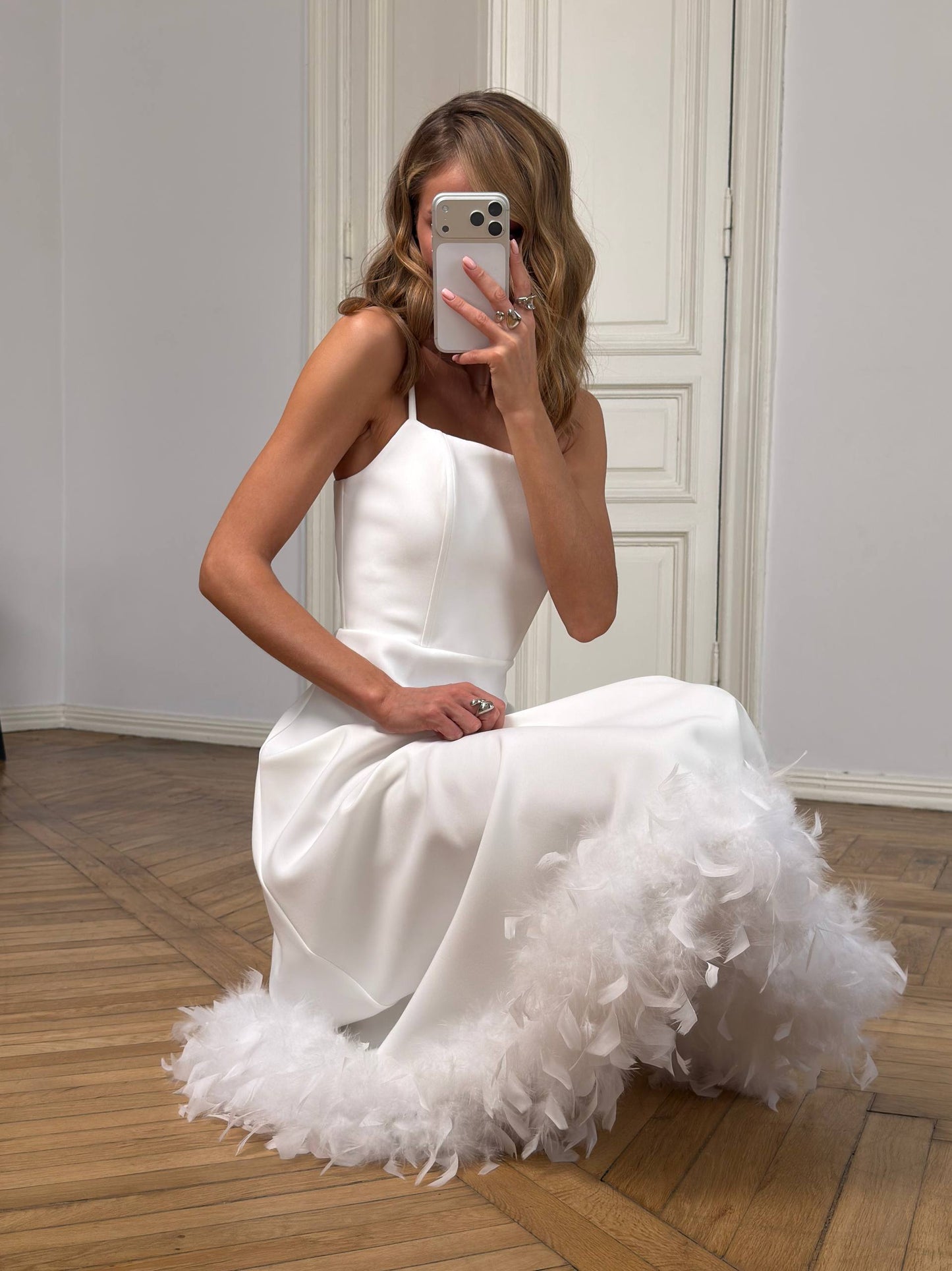 Woman in a white dress with a feathered skirt taking a mirror selfie.