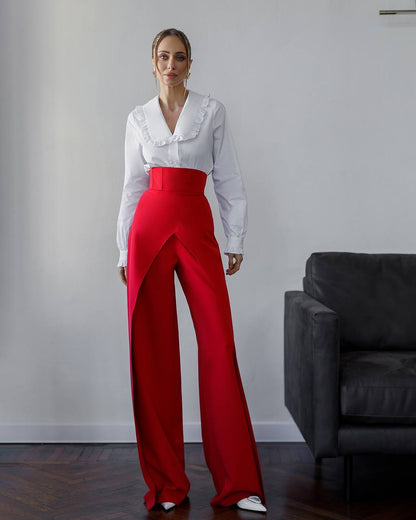 Woman wearing a white blouse and red pants standing in a room with a gray wall and black chair.
