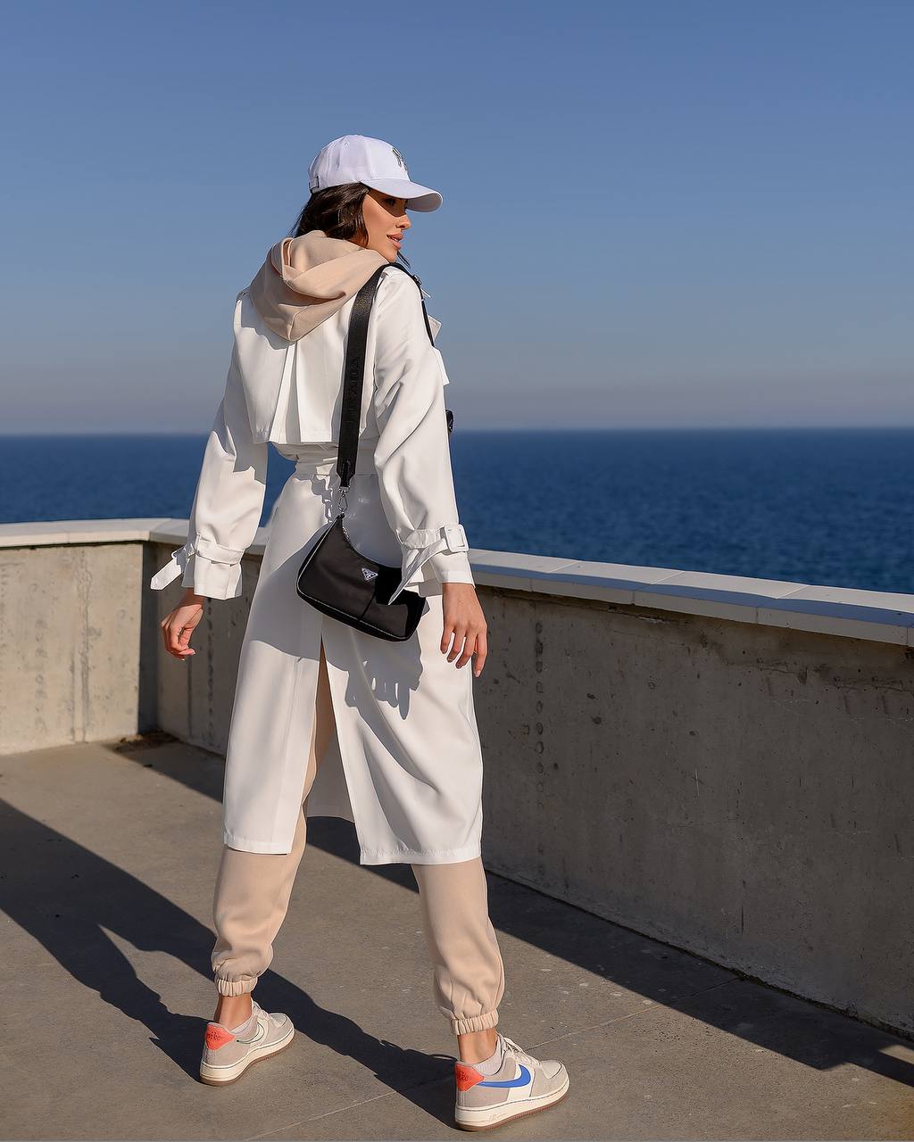 Person wearing a white outfit with a black bag, standing on a rooftop with a blue sky and ocean background.
