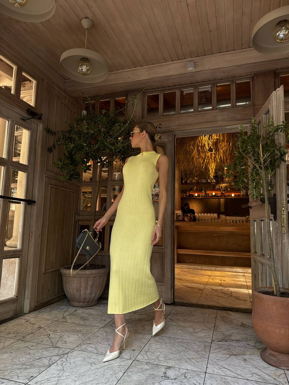 Woman in a yellow dress standing in a rustic interior setting with wooden walls and plants.