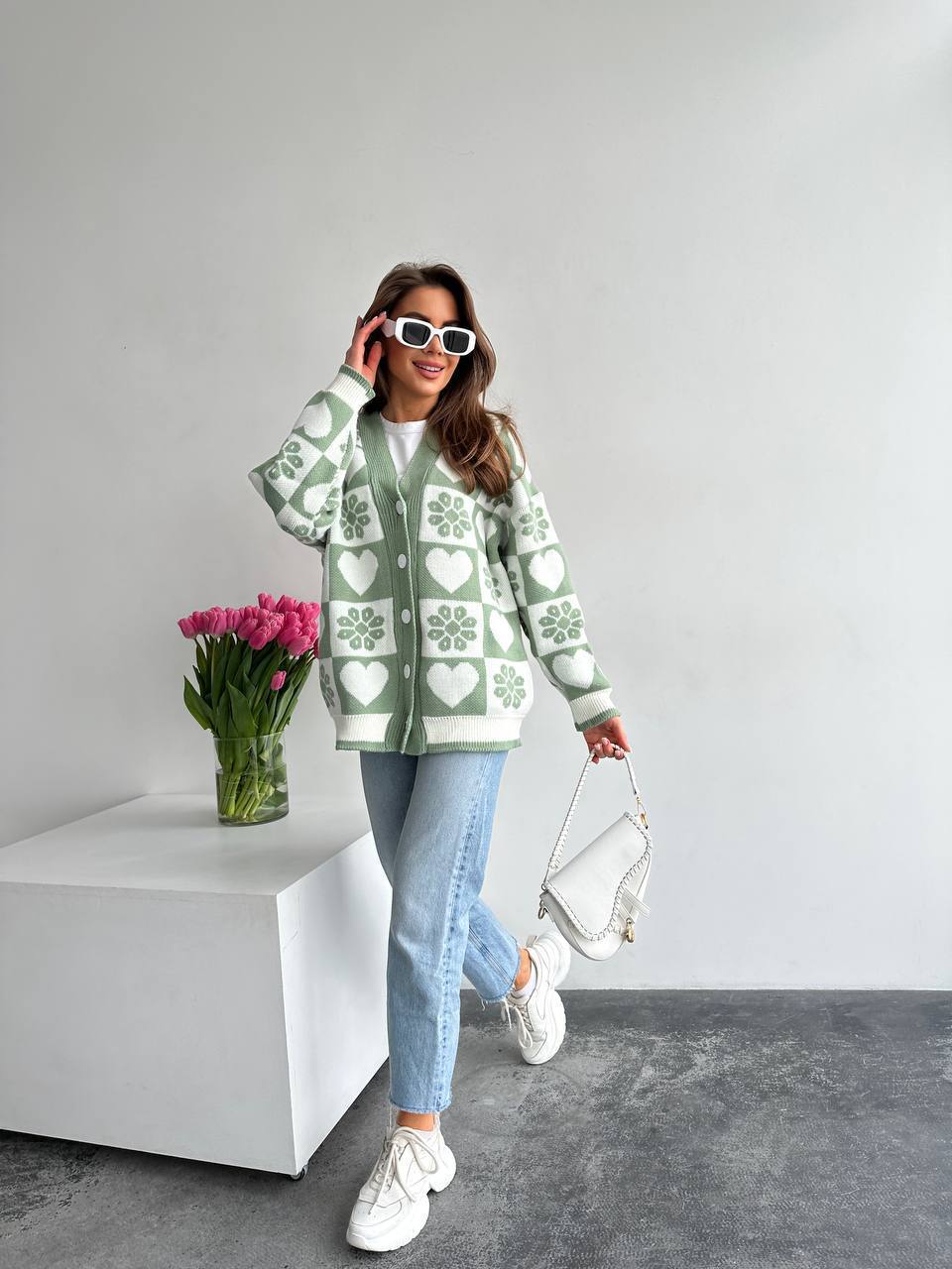 Woman wearing a green patterned cardigan with a vase of pink tulips on a white surface.