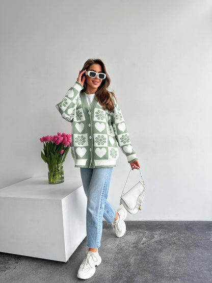 Woman wearing a green patterned cardigan with a vase of pink tulips on a white surface.