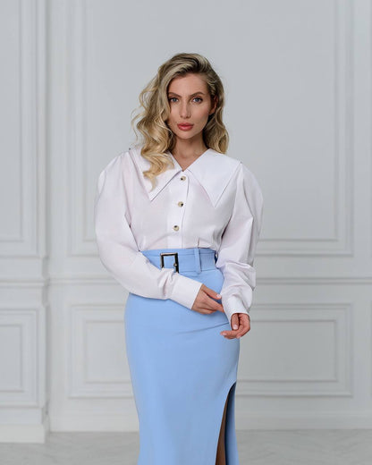 Woman wearing a white blouse and light blue skirt against a white wall.
