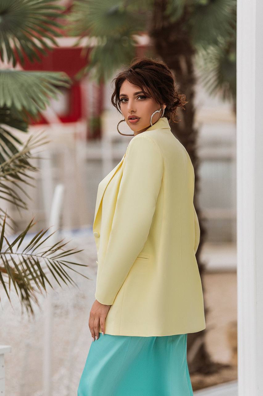 A woman wearing a pale yellow blazer and a turquoise skirt, photographed outdoors with palm leaves and a blurred architectural background.