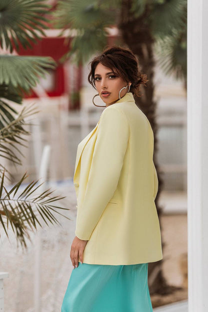 A woman wearing a pale yellow blazer and a turquoise skirt, photographed outdoors with palm leaves and a blurred architectural background.