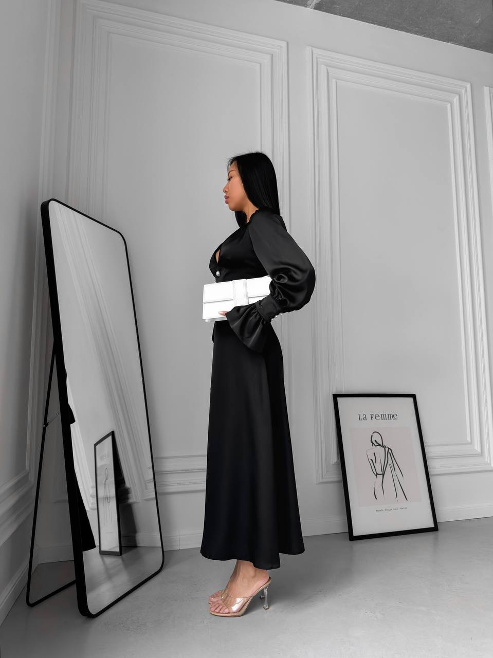 Woman in a black dress standing next to a mirror in a room with white walls and a framed picture.
