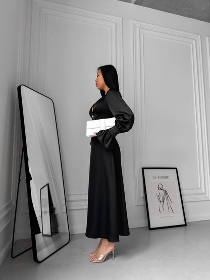 Woman in a black dress standing next to a mirror in a room with white walls and a framed picture.