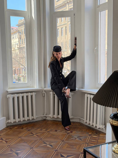 Person in formal attire standing in a room with large windows and a lamp.